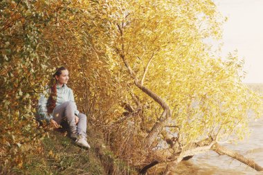 Genç bir kadın sonbahar yapraklarıyla nehir kıyısında oturuyor..