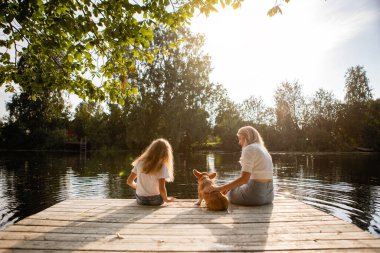 Kızıyla birlikte bir kadın ve safkan bir corgi pembroke köpeği gölün kıyısındaki ahşap iskelede oturuyor. Arka plan.