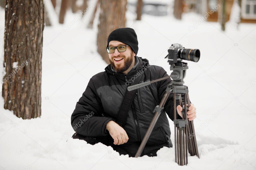Videographer makes a video filming a camera on a tripod. Bearded bespectacled man in warm clothes in a snowy park in winter.