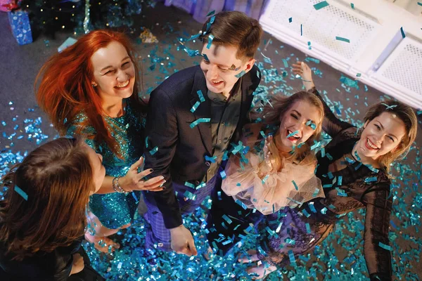 Group of friends celebrates the New Year with confetti near the Christmas tree. People laugh and rejoice.