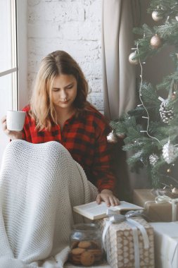Sarışın genç kız Noel ağacının yanında pencerenin yanında oturuyor. Kırmızı ekose gömlek, uzun bol saç, sıcak ekose, hediye kutuları ve bir fincan çay veya kahve. Seçici odak.