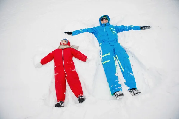 Snow Angel. Dad and son in winter red and blue clothes lie in the snow. Selective focus, blurred background.