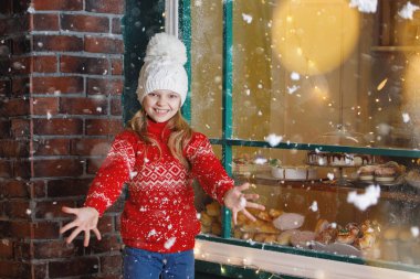 Noel kahvecisi yemek yapıyor. Beyaz örgü şapkalı sarışın bir genç kızın portresi donutlarla dolu bir gösterinin arka planında seviniyor. Seçici odaklanma. Bulanık ışıklar.