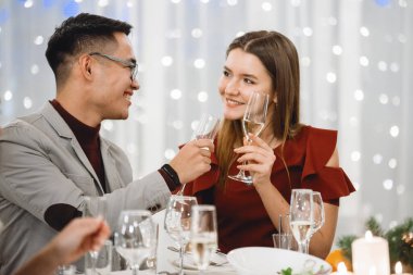 Şenlik masasında şampanya kadehleriyle gülümseyen, restoranda konuşan genç bir çift. Seçici odak, bulanık ışıklar.