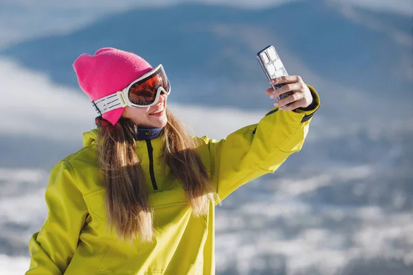 Woman in ski clothes takes a selfie against the backdrop of blue mountains. Yellow or mustard jacket with a hood, a knitted hat, ski goggles. Healthy lifestyle. Sports concept. Selective focus.