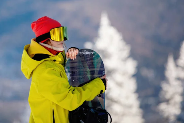 Sports concept. Woman snowboarder in ski clothing poses with a board against a background of blue mountains. Yellow jacket with a hood, knitted hat, ski goggles. Healthy lifestyle. Selective focus.