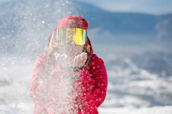 女性スキーヤーは手のひらから雪を吹く 青い山の斜面 赤いフード付きジャケット 黄色のスキーゴーグル 健康的なライフスタイル スポーツの概念 選択的焦点 — ストック写真