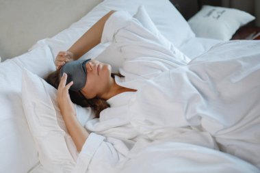 Yüzünde uyku maskesiyle beyaz bir yatakta uyuyan güzel bir genç kadın. Sağlıklı dinlenme.