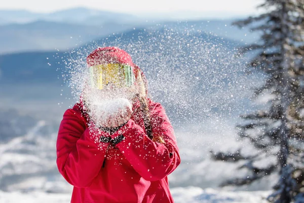 女性スキーヤーは手のひらから雪を吹く 青い山の斜面 赤いフード付きジャケット 黄色のスキーゴーグル 健康的なライフスタイル スポーツの概念 選択的焦点 — ストック写真