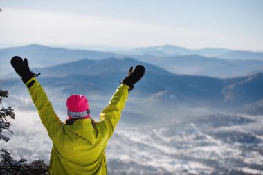 Kayak ekipmanlı kadın, uzak mavi dağların ve çukurların arka planında poz veriyor. Sarı ya da kapüşonlu hardal ceketli, örgü şapkalı. Arkadan bak. Sağlıklı bir yaşam tarzı. Spor konsepti. Seçici odak.