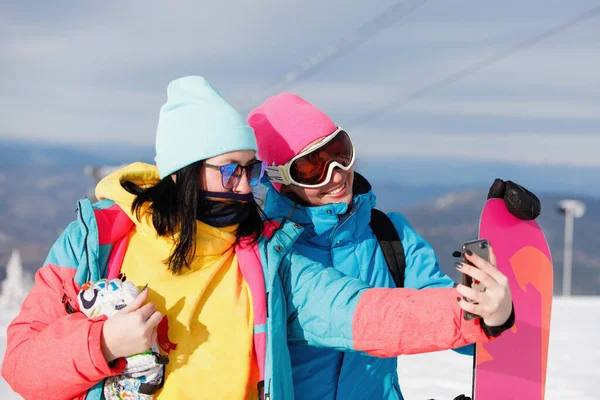 Women in ski clothes takes a selfie against the backdrop of blue mountains. Multicolor jacket with a hood, a knitted hat, ski goggles. Healthy lifestyle. Sports concept. Selective focus.