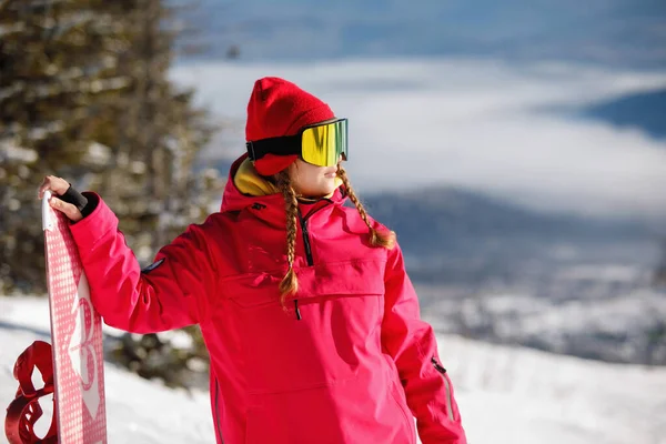 スキーウェアの女性スノーボーダーは 青い山々とスプルースを背景にボードでポーズをとっています フード付きジャケット 赤いパンツ スキーゴーグル 健康的なライフスタイル スポーツの概念 選択的焦点 — ストック写真