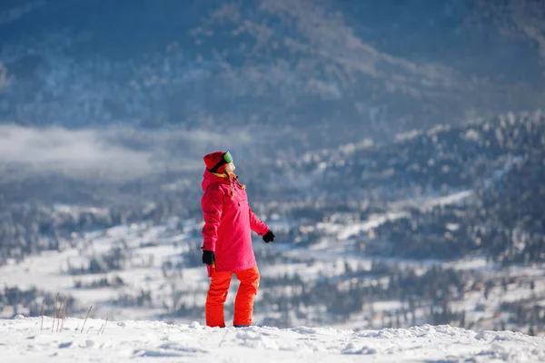 スキー用具の女性は青い山脈を背景にポーズをとっています フード付きジャケット 赤いパンツ スキーゴーグル 健康的なライフスタイル スポーツの概念 選択的焦点 — ストック写真