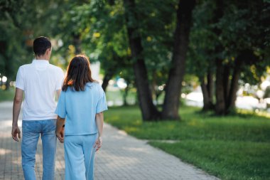 Adam ve kadın el ele tutuşur, yaz parkında yürür. Arkadan bak. Seçici odak, bulanık arkaplan.