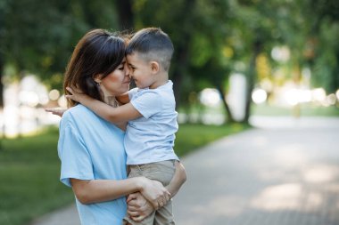 Anne yaz parkında oğluna sarılıp öpüyor. Seçici odaklanma, bulanık arkaplan. Mutlu bir çocukluk. Ebeveyn bakımı.