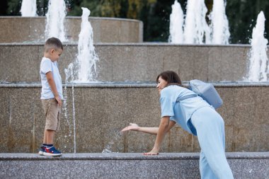 Anne ve oğlu yaz parkında çeşmenin yanında. Seçici odaklanma, bulanık arkaplan. Mutlu bir çocukluk. Ebeveyn bakımı.