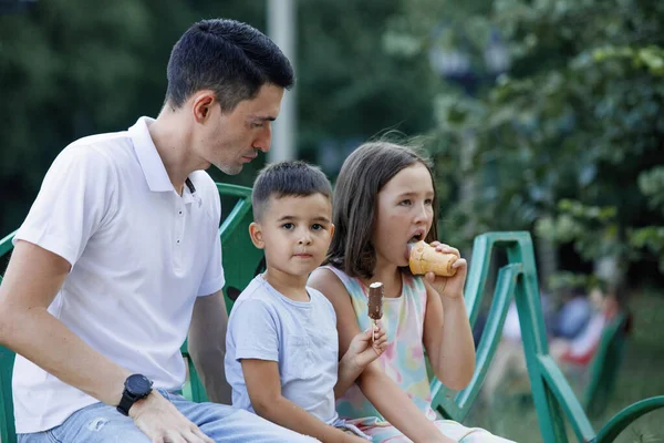 Daddy with daughter and son eating ice cream on a bench in the summer park. Selective focus, blurred background. Happy childhood.