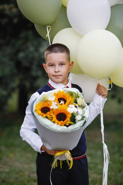 Boy in school uniform with flowers and balloons posing in a summer park. Selective focus, blurred background. Primary School.