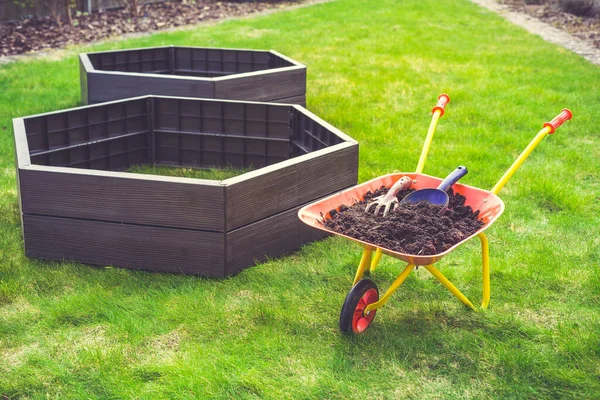 Garden barrow with soil and empty raised beds on grass prepared for filling with soil. Urban gardening concept.