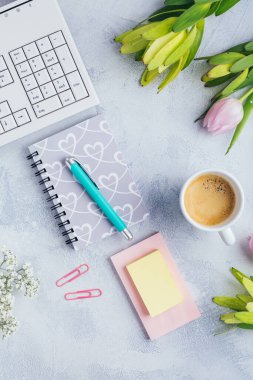 Bir fincan kahve, boş bir defter, klavye, malzeme ve çiçeklerle dolu bir işyeri.