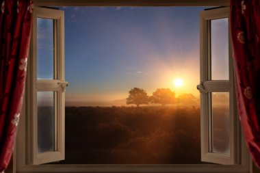 Perdeleri olan, birkaç ağaçla birlikte kırsal bir arazide altın bir güneşin doğuşunu seyreden dul bir kadın. Şafağın ilk ışıklarında parlayan güneş ışığı ve sensörler.