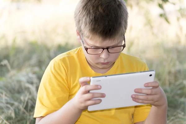 Chlapec v brýlích vypadá tabletový počítač v přírodě — Stock fotografie