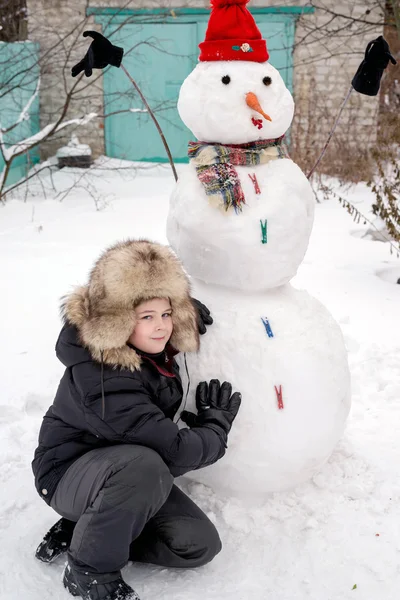 Pojken i en pälsmössa runt snögubbe — Stockfoto