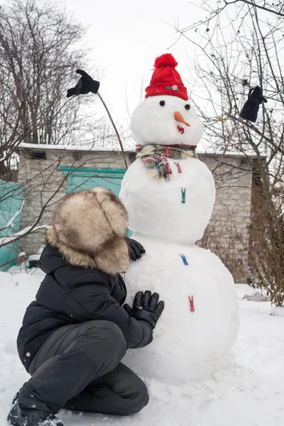 Der Junge mit der Pelzmütze um den Schneemann — Stockfoto