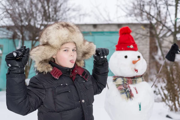 O rapaz com um chapéu de peles em volta do boneco de neve — Fotografia de Stock