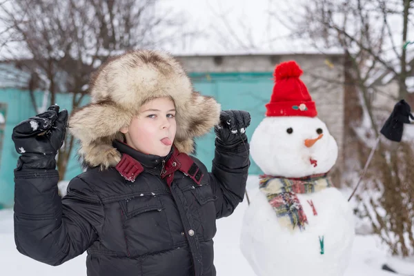 Il ragazzo con un cappello di pelliccia intorno a pupazzo di neve — Foto Stock