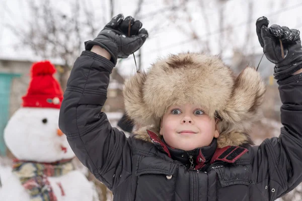 Il ragazzo con un cappello di pelliccia intorno a pupazzo di neve — Foto Stock