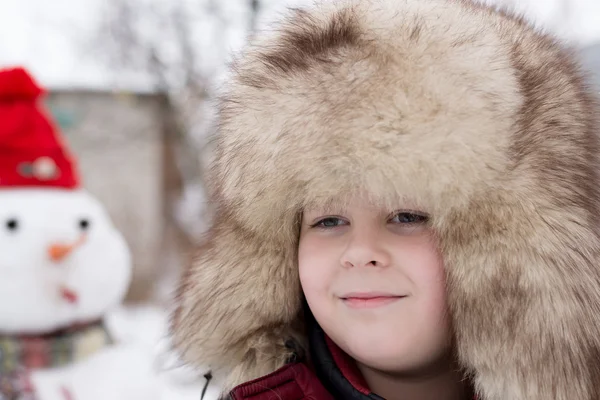 Мальчик в меховой шапке рядом со снеговиком — стоковое фото