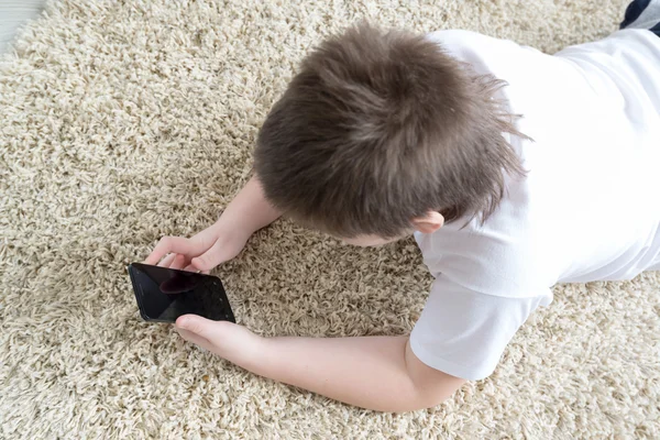 Jongen met telefoon liggend op een tapijt in de kamer — Stockfoto