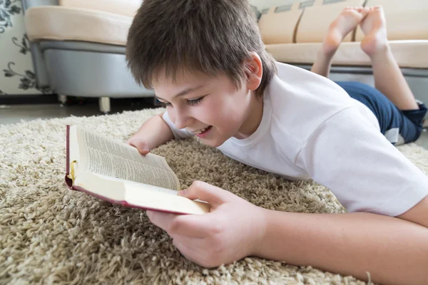 Pojke läser en bok liggande på matta i rummet — Stockfoto