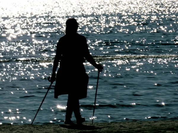 Silhouette of a woman with sticks walking along the seashore in the rays of the sun reflecting from the water
