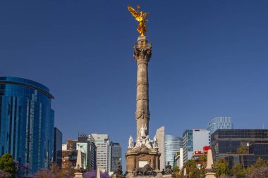 Mexico City, Meksika - 14 Mart 2022: Güneşli bir bahar gününde Özgürlük Anıtı ve döner kavşak, modern binalar ve arka planda çiçek açan Jacaranda ağaçları