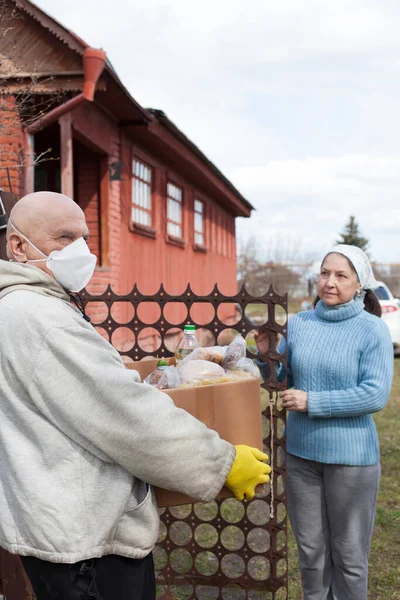 Senior male volunteer in mask gives an elderly woman boxes with food near her house. Husband  helps his elderly wife. Family support, caring. Quarantined, isolated. Coronavirus covid-19. Donation