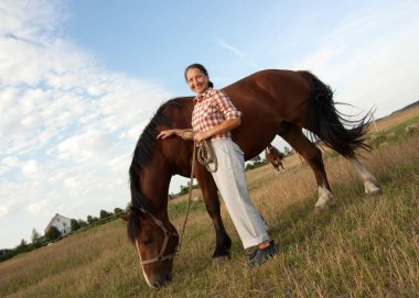 Güzel, olgun bir kadın yüzünde mutlu bir gülümsemeyle atın yanında duruyor.