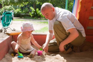 Mutlu baba ve yürümeye başlayan çocuk kum havuzunda kumla oynuyorlar.