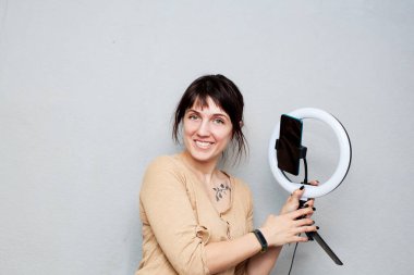 Köprücük kemiğinde dövmesi olan kız akıllı telefondan video kaydediyor ve gri arka planda yüzük lambasıyla kendini aydınlatıyor.