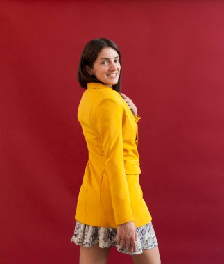 Rusya 'dan genç bir kadın kırmızı arka planda sarı ceketli poz veriyor, omzunun arkasından arkasını dönüyor.