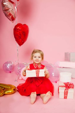  Pembe arka planda kırmızı elbiseli, hediyeli ve balonlu üç yaşında bir kız..