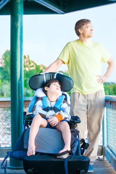 Padre sentado junto al lago con su hijo discapacitado en silla de ruedas — Foto de Stock