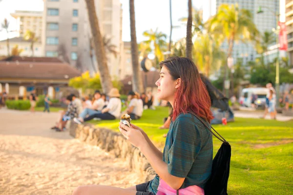 若いです女の子座っていますでワイキキビーチ近くの夕日食べるスパムMusbiとともに群衆の観光客が背景にぼやけて — ストック写真