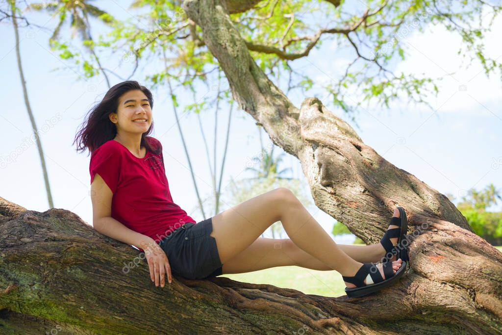 Biracial teen girl relaxing in large tree branch at park by ocean on island of Oahu, Hawaii