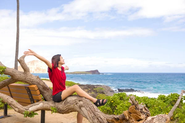 ハワイ州オアフ島のMakapu Uビーチパークで熱帯の海によって岩のビーチで倒木の幹に座っている赤いシャツの10代の女の子 — ストック写真