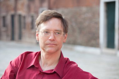 İtalya 'da kırmızı tişörtlü beyaz bir adam kameranın karşısına bakıyor. Venedik' in tarihi tuğla binaları arka planda.