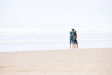 Pasifik Okyanusu kıyısında sisli bir günde öpüşen melez genç çift, pantolonları kıvrılmış.