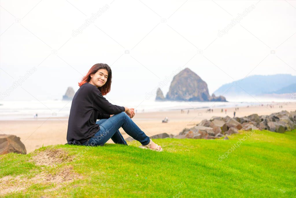 Biracial teen girl sitting on grassy knoll or hill by shore near the ocean in the Pacific Northwest on foggy day