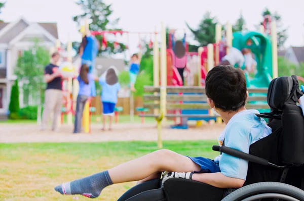 Behinderter kleiner Junge im Rollstuhl sieht Kindern beim Spielen zu — Stockfoto